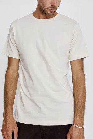 A man with light skin wears the Mr Simple Reginald Organic Tee in off-white—an easy-fitting, midweight t-shirt—paired with dark pants. Only his torso and lower face appear; a silver chain bracelet is on his right wrist.