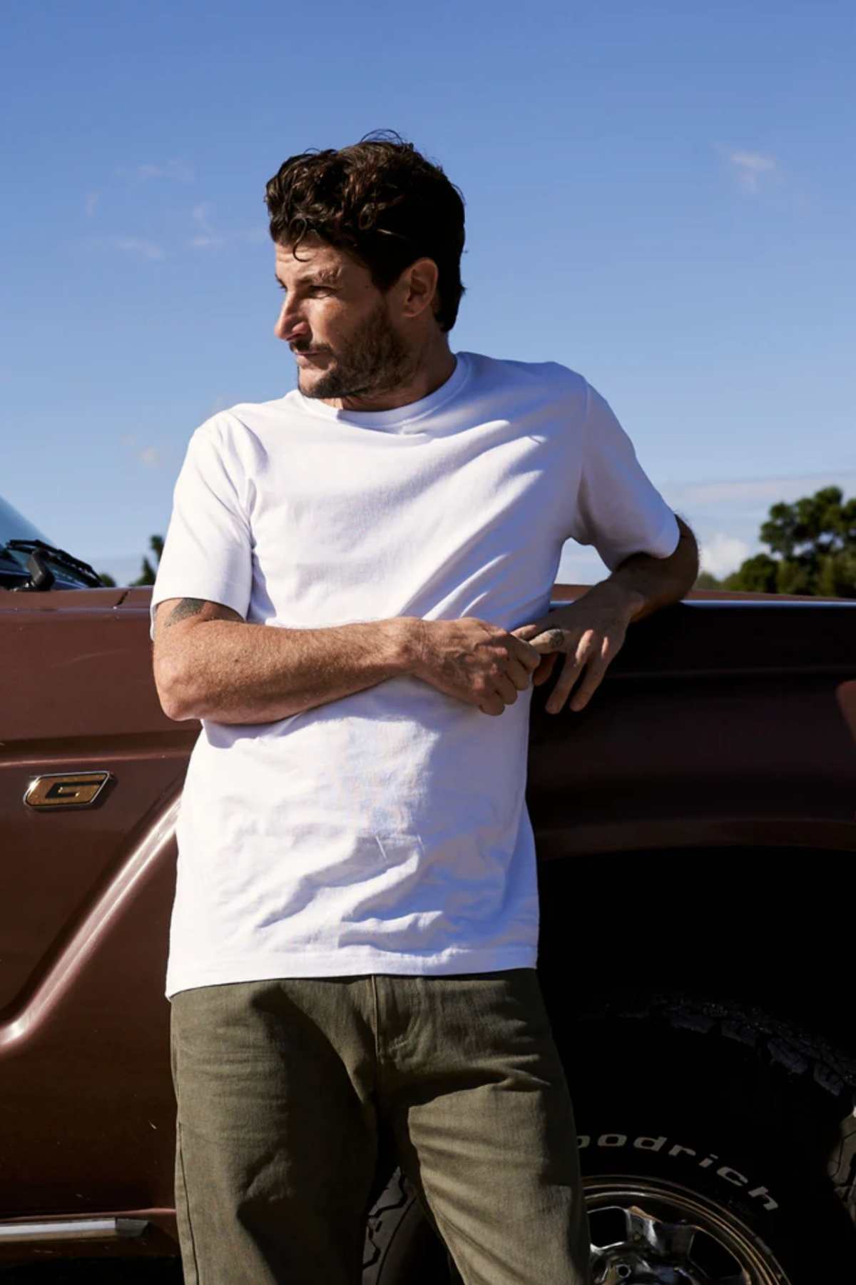 A man in the Mr Simple Reginald Organic Tee and olive green pants leans casually against a dark brown truck, squinting to his left beneath a clear blue sky.