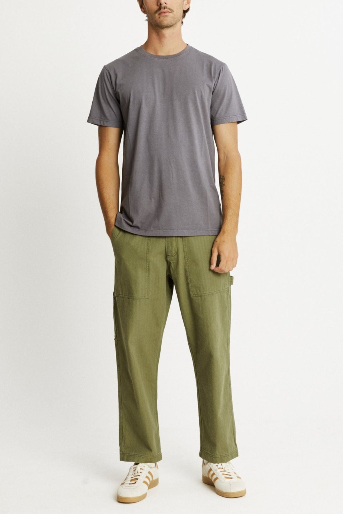 Dressed in the Mr Simple Reginald Organic Tee against a plain white backdrop, an individual sports gray attire complemented by olive green cargo pants and white sneakers. Their left arm resting calmly at their side adds to the relaxed aura, highlighting the stretchy and durable design of their ensemble.
