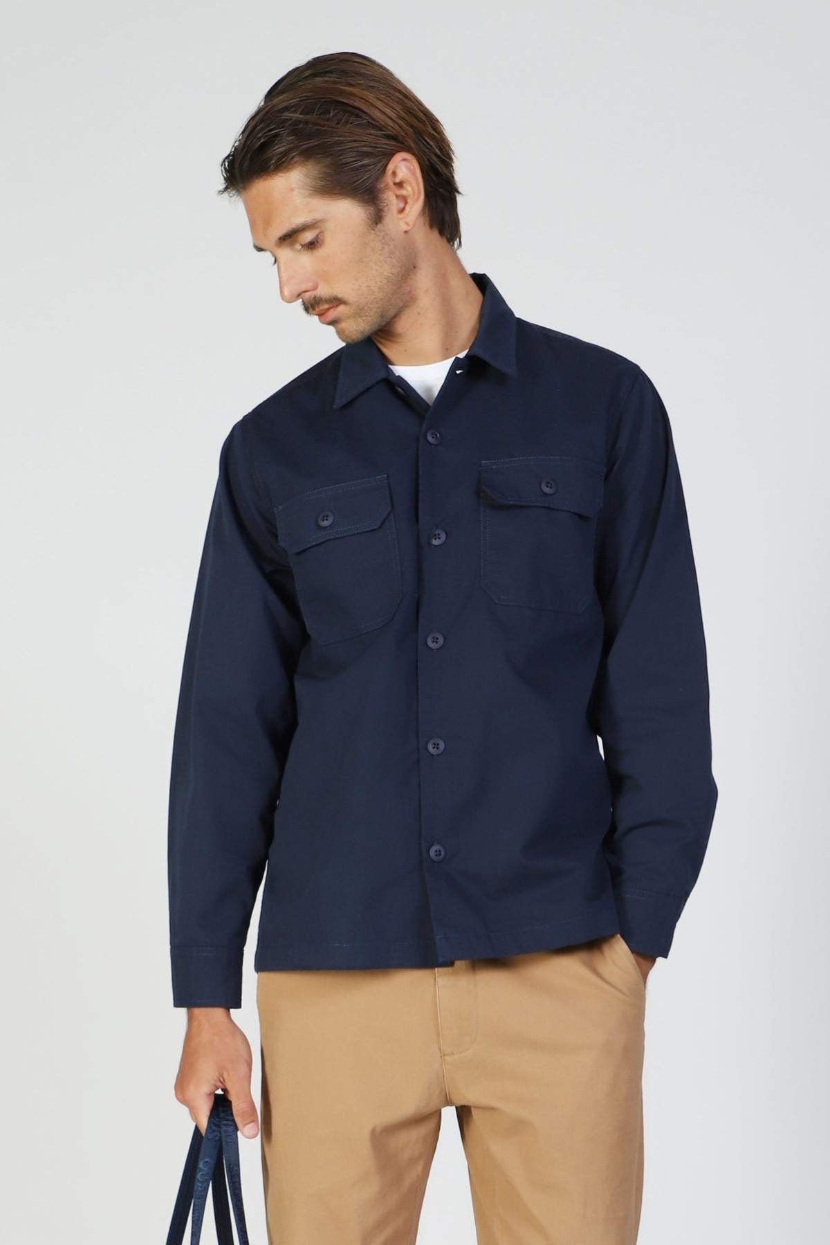 A man wears the Mr Simple Ripstop Canvas Overshirt with chest pockets and tan pants, standing against a plain light background and holding a navy bag by his side as he looks down.