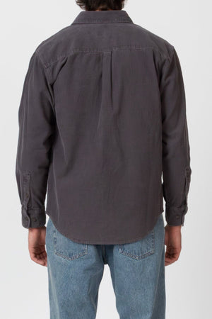 A person faces away from the camera, wearing the Mr Simple Sawyer Cord Shirt in dark gray with light blue jeans. The background is plain and light-colored.