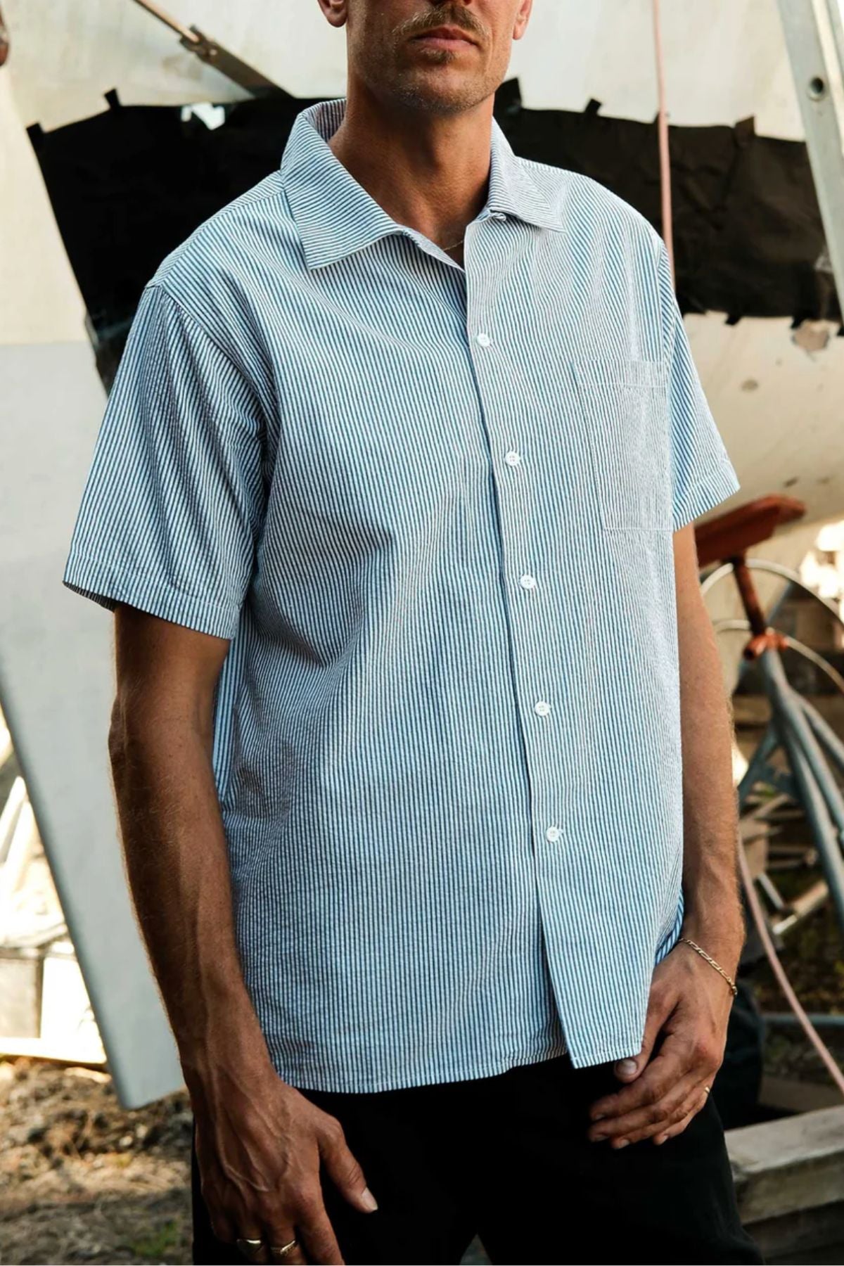 A man, partially out of frame, stands outdoors wearing the Mr Simple Seersucker Short Sleeve Shirt in light blue and white stripes. Behind him are construction materials and a partially built structure.
