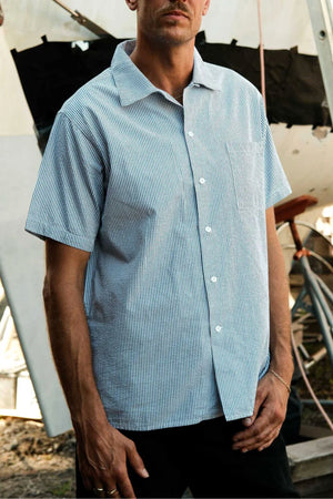 A man, partially out of frame, stands outdoors wearing the Mr Simple Seersucker Short Sleeve Shirt in light blue and white stripes. Behind him are construction materials and a partially built structure.