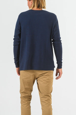 A person with shoulder-length blonde hair stands with their back to the camera, wearing a navy Mr Simple Smith Waffle long sleeve shirt and tan pants against a plain white background.