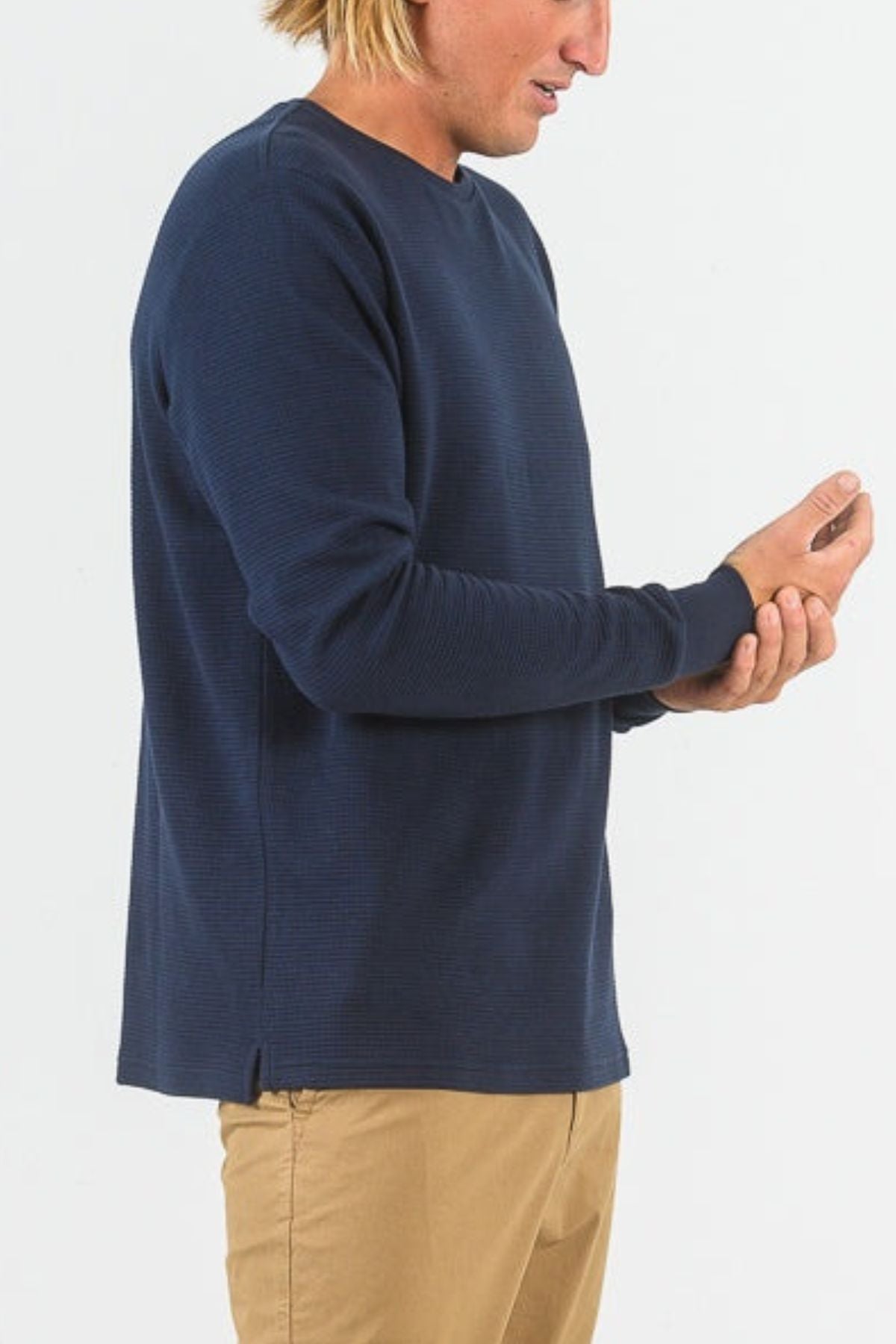 A person with blonde hair wearing a navy blue Mr Simple Smith Waffle long sleeve shirt and khaki pants stands against a plain white background, looking at and holding their left hand.