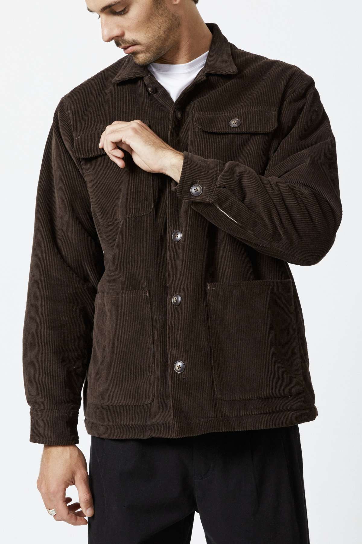 The man in a Mr Simple Stockyard Sherpa Jacket, featuring a full button front and two large pockets, adjusts a button while looking down. His short hair complements classic black pants against a plain white background, emphasizing the timeless style of his outfit.