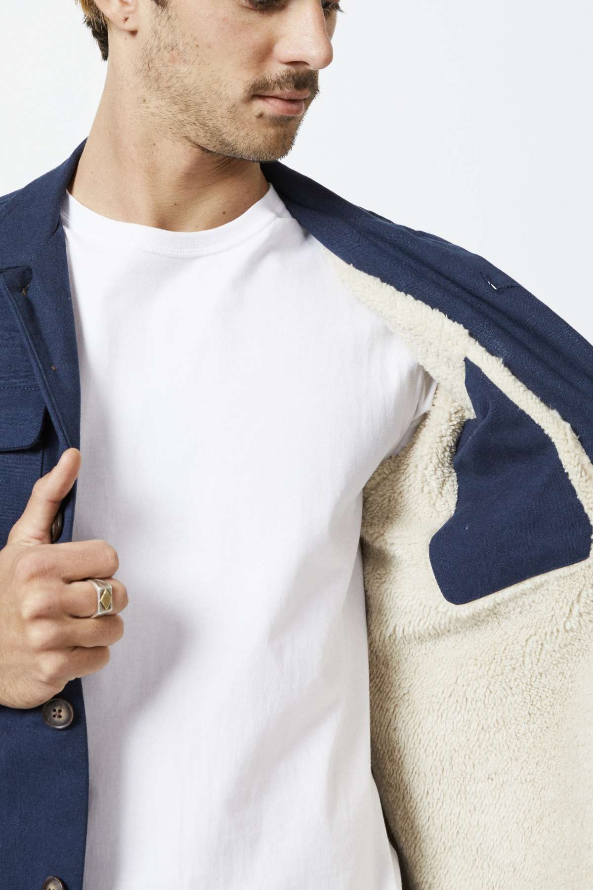 A man is highlighting the interior of a Mr Simple Stockyard Sherpa Jacket. The blue jacket boasts a plush, cream-colored sherpa lining. He uses his hand to hold one side open, unveiling a plain white t-shirt underneath and showcasing a ring on his middle finger. His face is partially visible against the uncomplicated white backdrop.