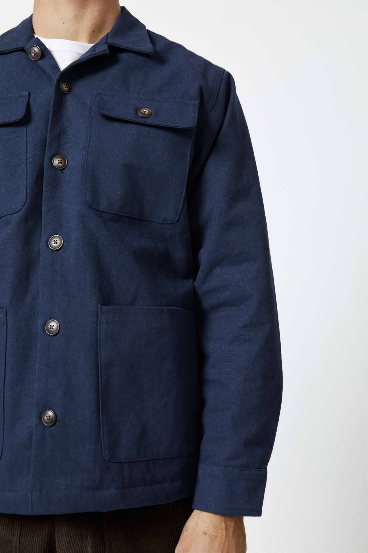 A person wearing a Mr Simple Stockyard Sherpa Jacket, made of 100% cotton in navy blue, with large front pockets and a button-up design. The jacket is buttoned up, highlighting its sherpa lining, as the person stands against a plain, light-colored background. The image focuses on the person's torso and arm.