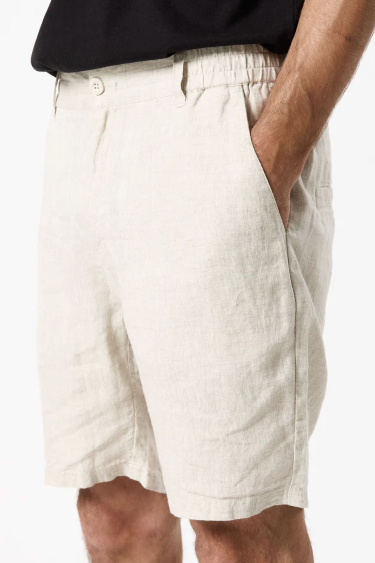 A person wears Mr Simple Tanner 3.0 Linen Shorts in breathable light beige with an elastic waistband and side pockets, one hand in their pocket, effortlessly paired with a black shirt.