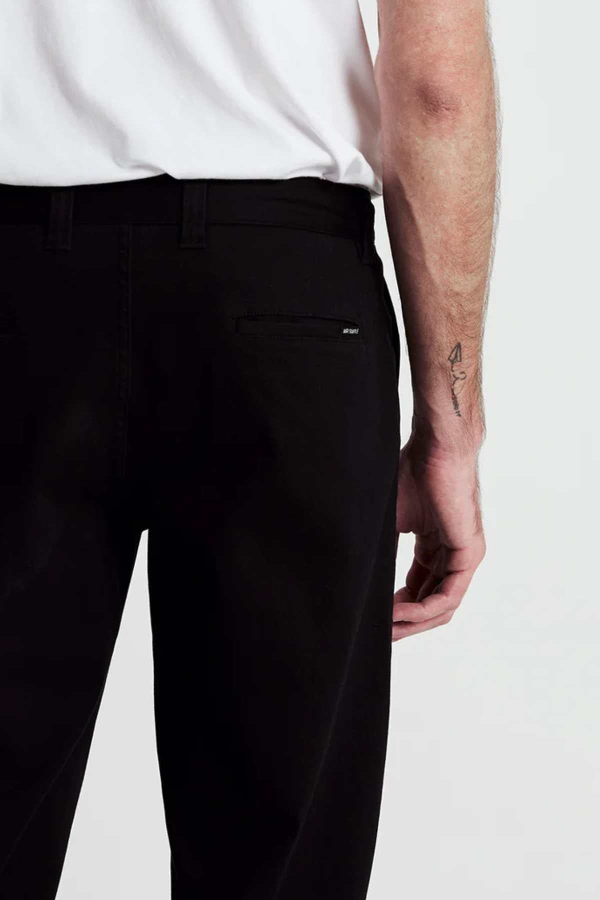 Someone shown from the waist down wears Mr Simple Wilson Standard Chino black pants and a white shirt. The pants feature a zippered back pocket, their right arm with a small forearm tattoo is visible.