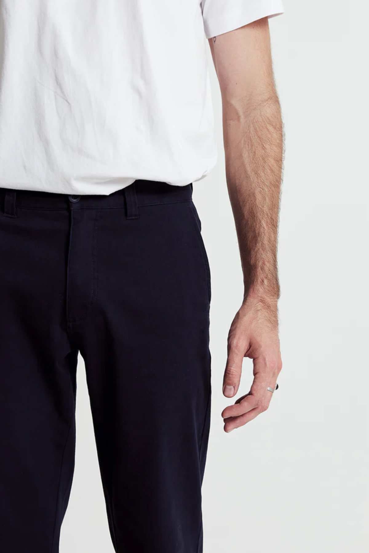 The image displays a person from the waist down wearing dark Mr Simple Wilson Standard Chino pants and a relaxed-fit organic cotton white t-shirt. Their right arm hangs at their side, showing a small forearm tattoo against the plain, neutral background that emphasizes the outfit's simplicity.