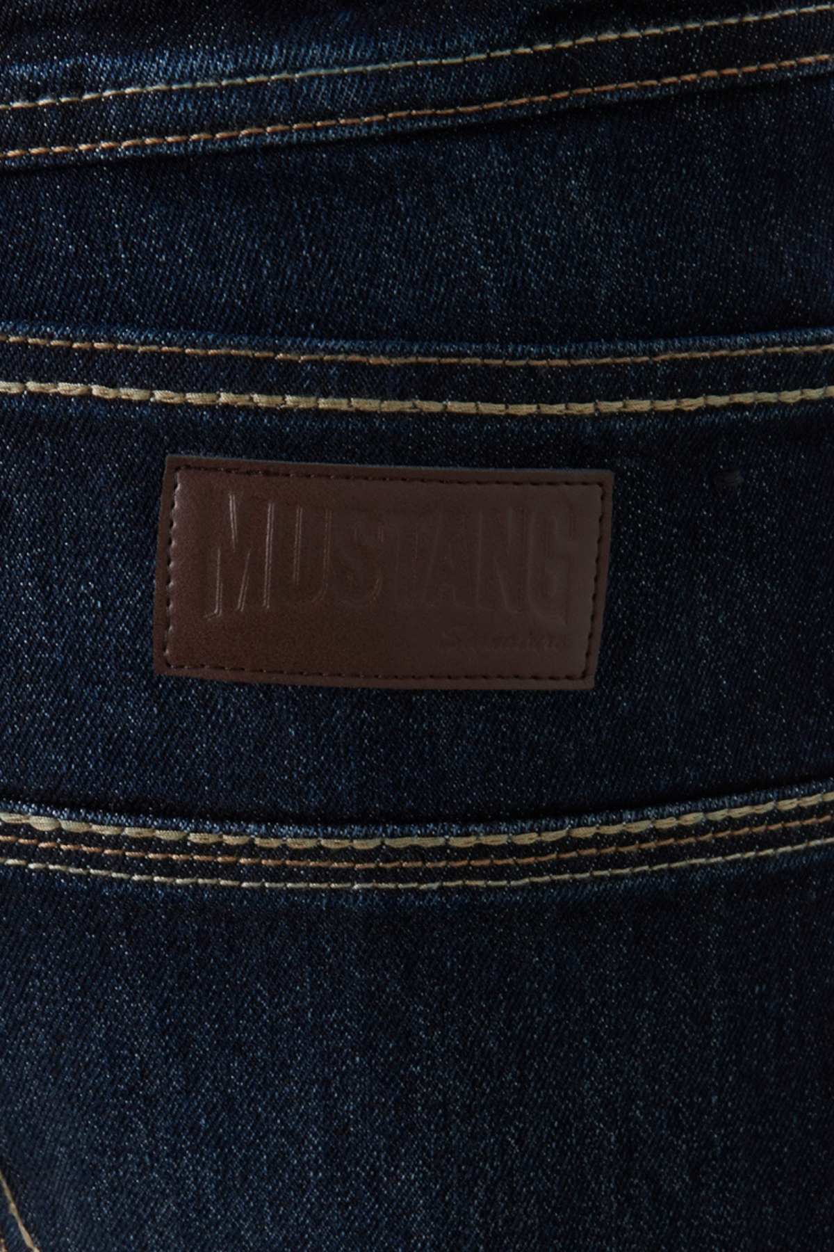 Close-up of the back pocket area of Mustang Signature Mens Jeans in dark blue denim, showing a brown leather patch embossed with "MUSTANG" and visible contrast stitching along the seams. Classic fit and premium quality.