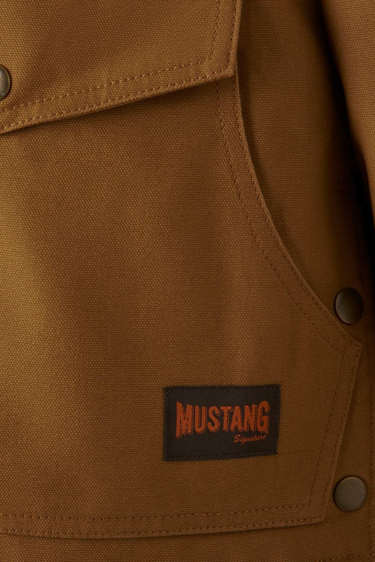 Close-up of a Mustang Signature Outback Canvas Jacket in brown, showcasing a pocket with four metallic buttons and a black label reading "Mustang Signature" in orange and beige text. The textured durable canvas fabric promises superior protection.