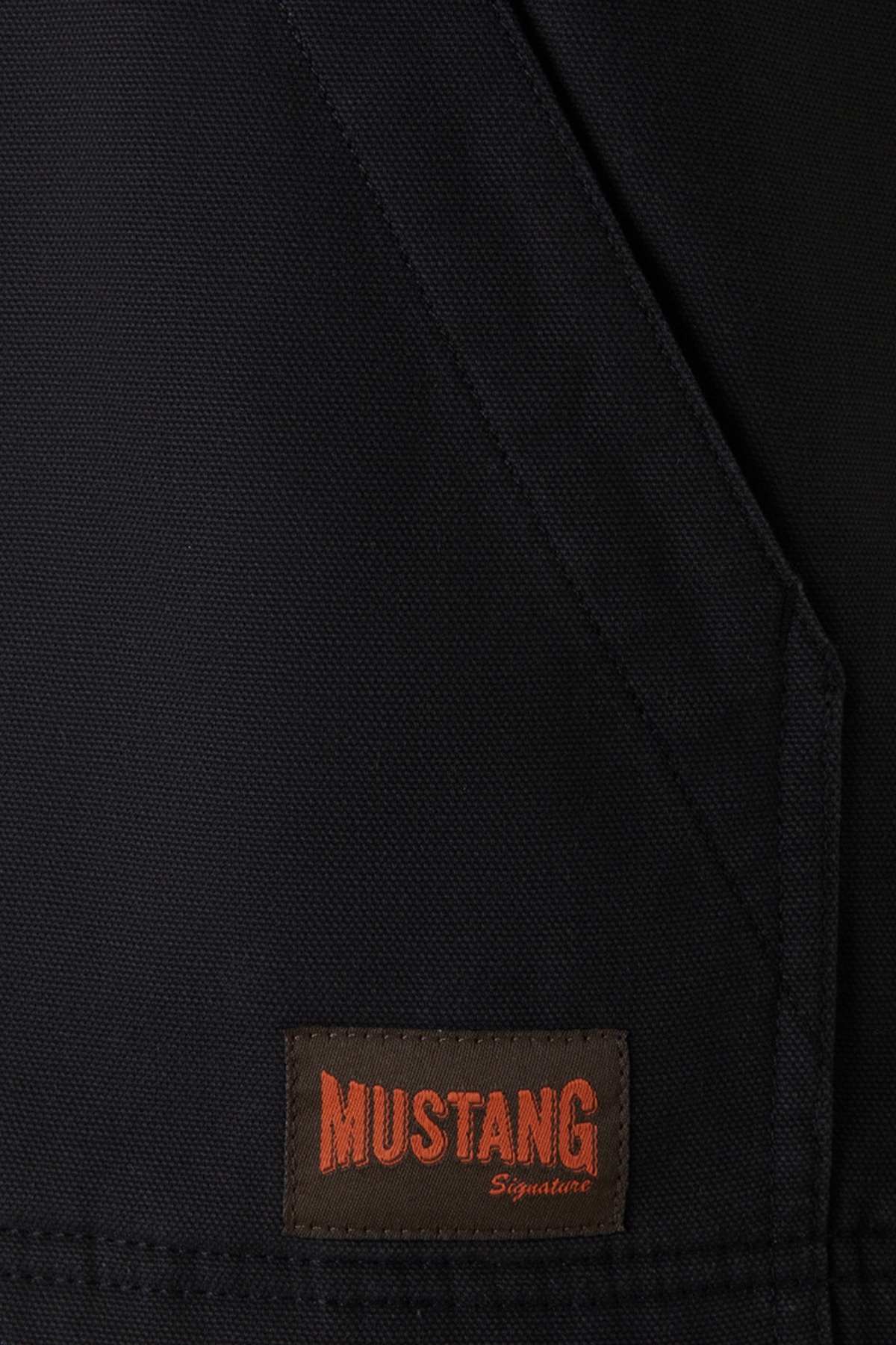 Close-up of a Mustang Signature Outback Canvas Vest made from durable canvas material, featuring a dark fabric with a brown patch label at the bottom. The label reads "MUSTANG Signature" in bold orange and cursive red lettering. The fabric has a seam running diagonally across the image, highlighting its functional features.