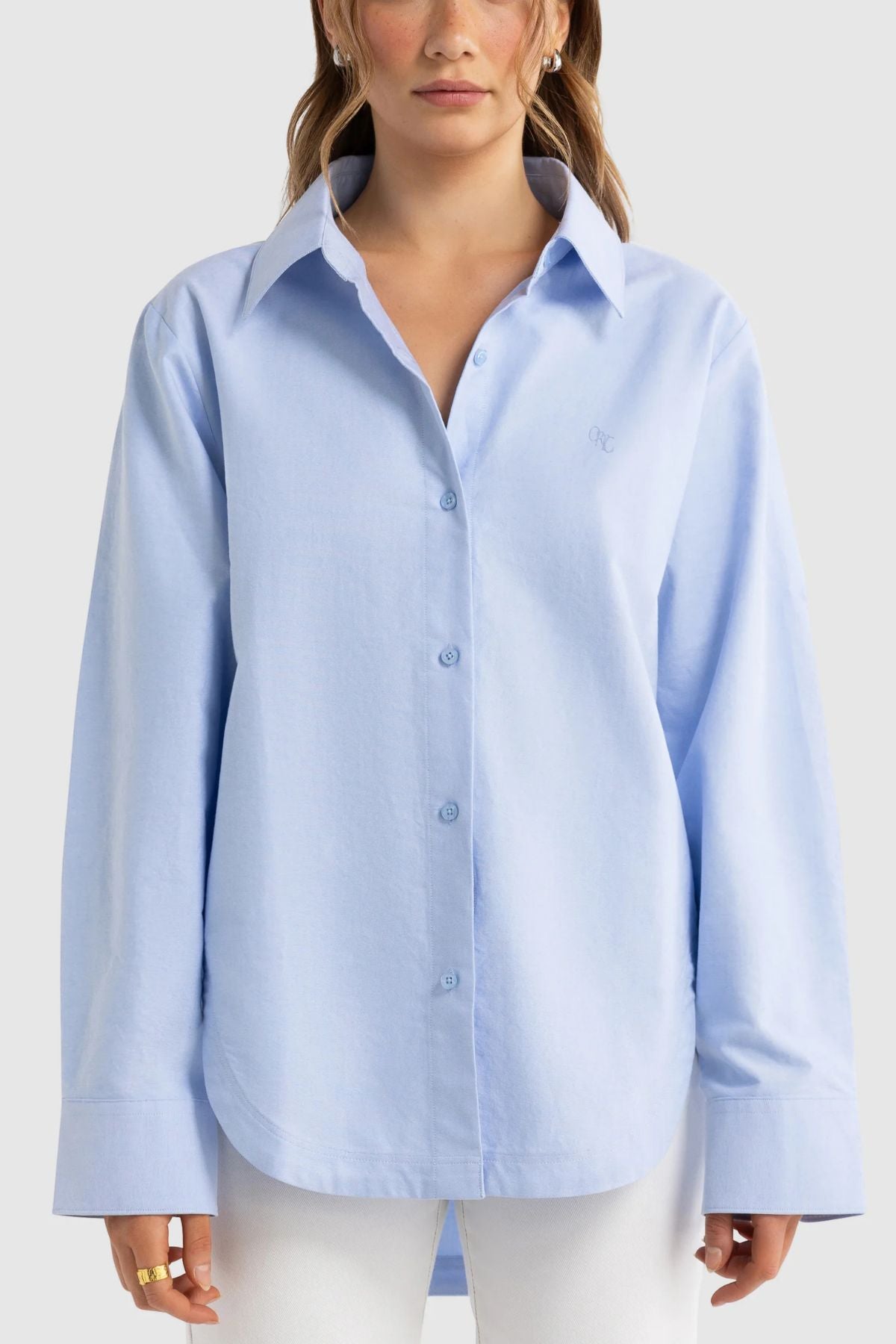 A woman wearing the ORTC Adelaide Oxford Shirt in an oversized fit and light blue color, featuring wide cuffs, paired with white pants stands against a plain light background. The premium cotton of the ORTC Adelaide Oxford Shirt enhances her relaxed and stylish appearance.
