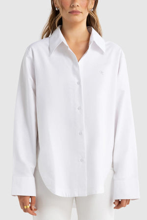 A person wearing the ORTC Adelaide Oxford Shirt in an oversized fit, featuring wide cuffs, paired with white pants and standing against a plain light gray background.