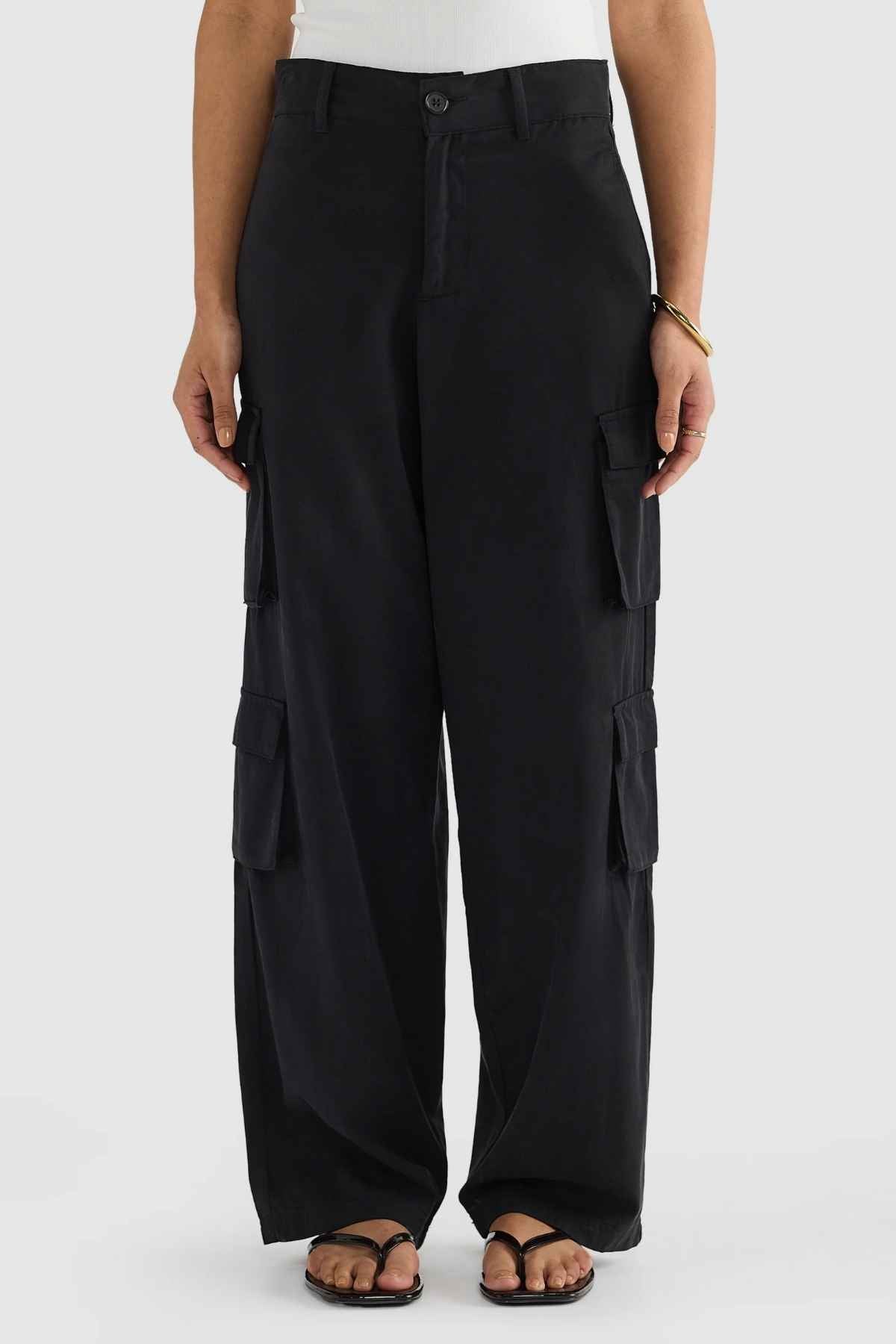 Someone stands on a light background wearing the ORTC Arden Cargo Pants in black, made from a tencel-cotton blend. These pants, featuring multiple cargo pockets, are paired with black flip-flops, a white top, and a bracelet is visible on their wrist.