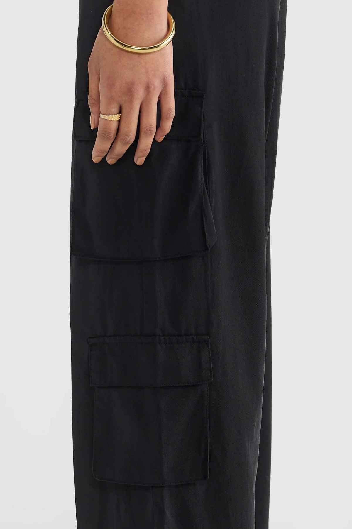 Dressed in ORTC Arden Cargo Pants crafted from a stylish tencel-cotton blend with two spacious cargo pockets, a person accessorized with a gold bracelet and ring poses against a light gray backdrop.