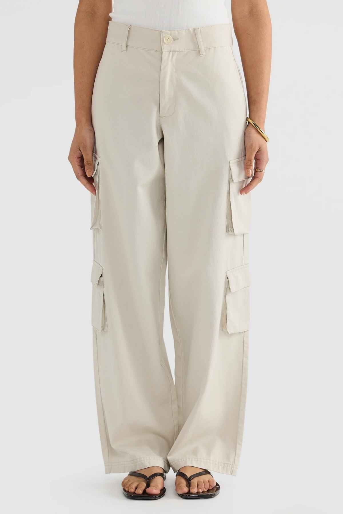 The person is wearing ORTC Arden Cargo Pants in a beige shade, featuring several cargo pockets. These pants are paired with a white top and black sandals. They stand in a neutral pose against a plain background, highlighting the seamless mix of tencel-cotton fabric in their outfit.