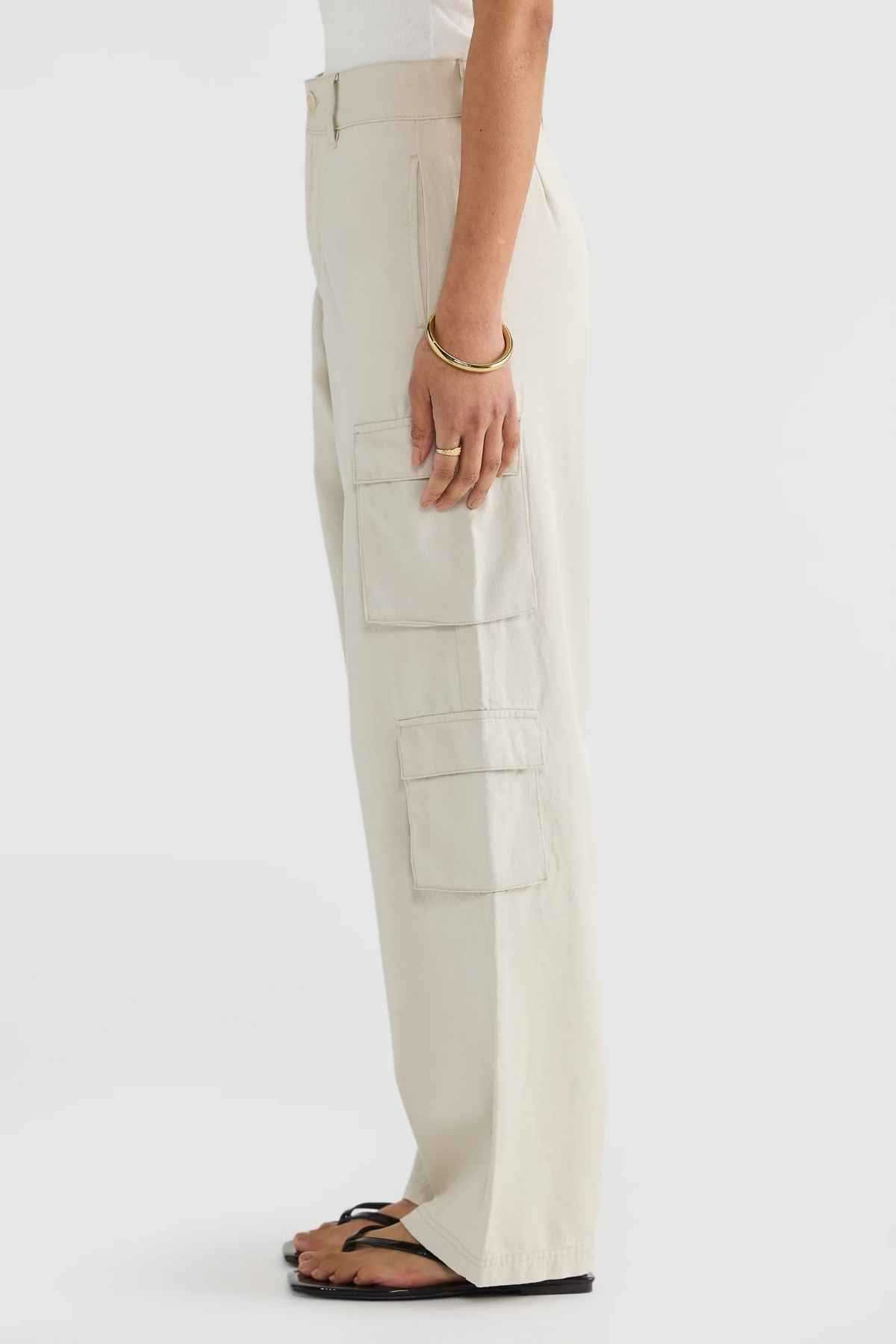 A person standing sideways is styled in distinctive ORTC Arden Cargo Pants, complemented by a crisp white top, black sandals, and a gold bracelet. The simple white background emphasizes the tencel-cotton blend of their outfit.