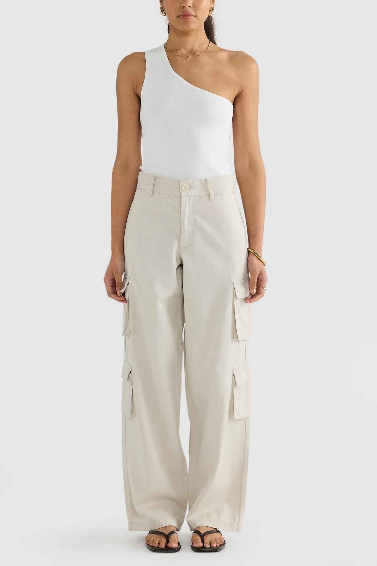 A person stands against a neutral background wearing the ORTC Arden Cargo Pants, crafted from a soft tencel-cotton blend in light beige. These stylish pants feature multiple cargo pockets and are paired with a chic white one-shoulder top and black sandals for a practical yet fashionable look.