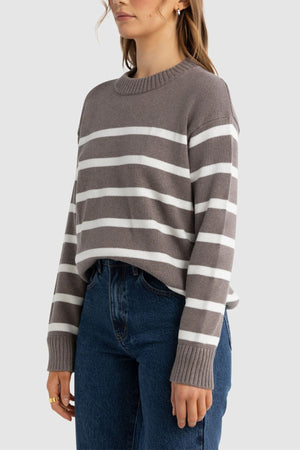 A woman wears the ORTC Astrid Crew Knit, a relaxed fit sweater with white horizontal stripes, paired with blue jeans and standing against a plain light gray background.