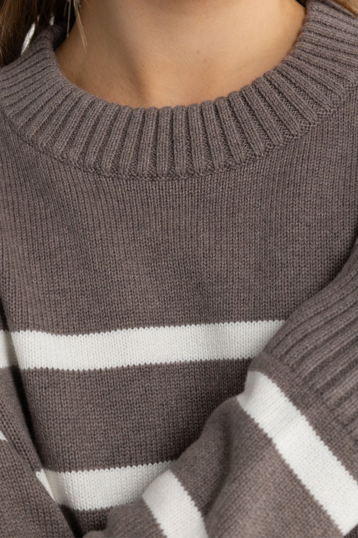 A close-up of a person wearing the ORTC Astrid Crew Knit, a luxury brown sweater with white horizontal stripes, ribbed neckline, relaxed fit, and detailed pattern.