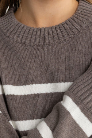 A close-up of a person wearing the ORTC Astrid Crew Knit, a luxury brown sweater with white horizontal stripes, ribbed neckline, relaxed fit, and detailed pattern.