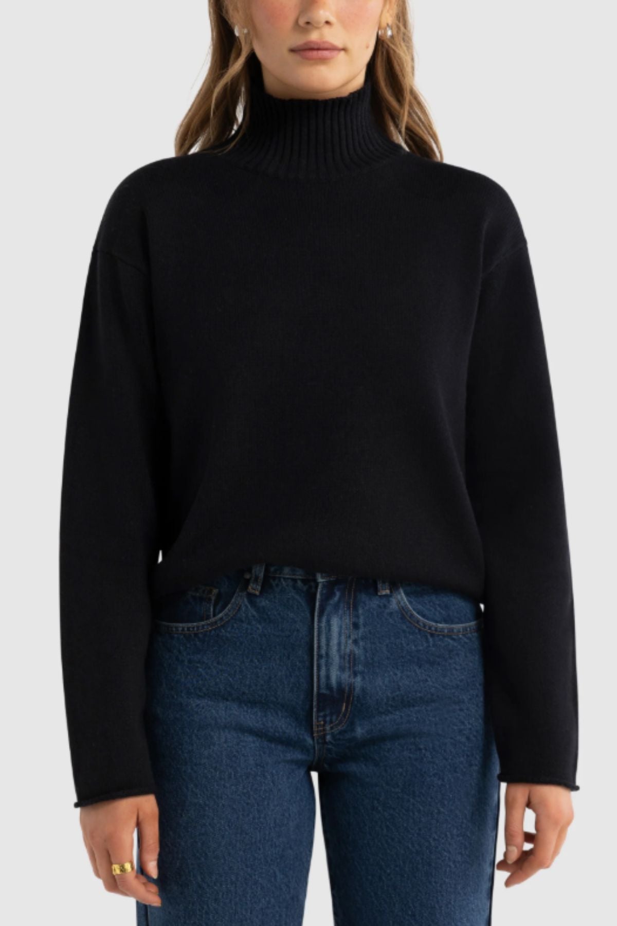 A woman stands facing forward against a light gray background, wearing the ORTC Astrid Knit Sweater in black and high-waisted blue jeans. Her hands are relaxed at her sides.