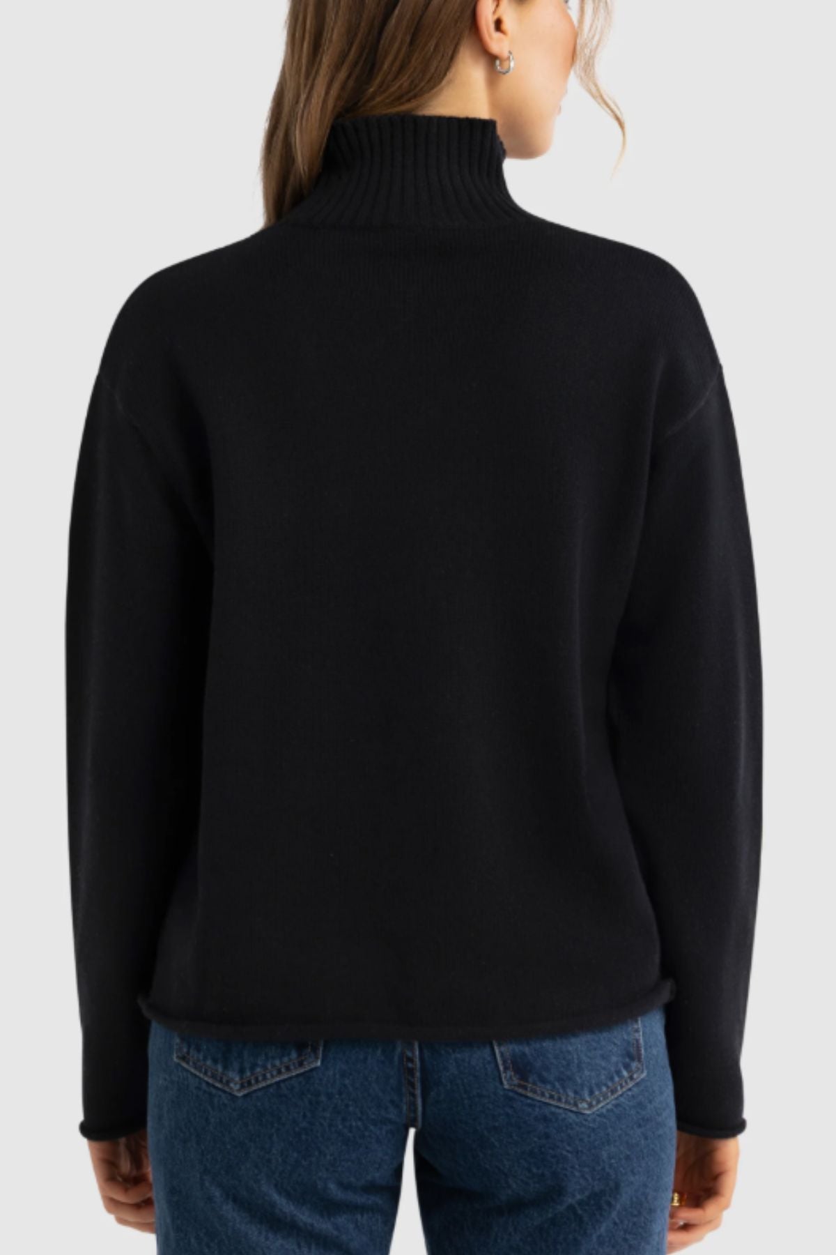 A woman is seen from the back, standing against a plain light gray background, wearing blue jeans and the ORTC Astrid Knit Sweater in black.