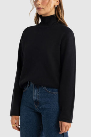 A woman with wavy brown hair, a gold hoop earring, and a ring wears the ORTC Astrid Knit Sweater tucked into high-waisted blue jeans against a plain background for an effortlessly chic look.