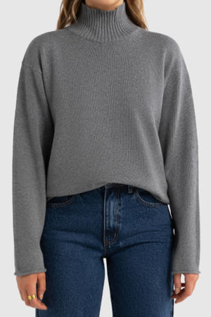 A person wearing the ORTC Astrid Knit Sweater in gray with long sleeves and blue high-waisted jeans stands against a light gray background.