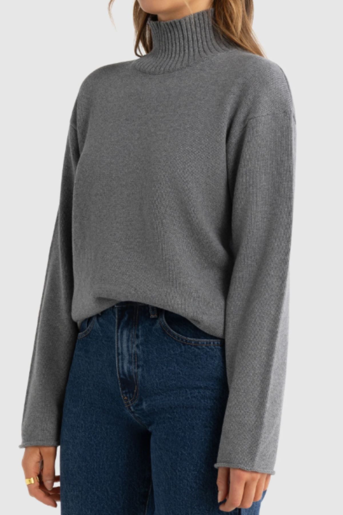 Someone wears the ORTC Astrid Knit Sweater—a gray cotton-cashmere women’s turtleneck with long sleeves—tucked into high-waisted blue jeans, standing against a plain light background.