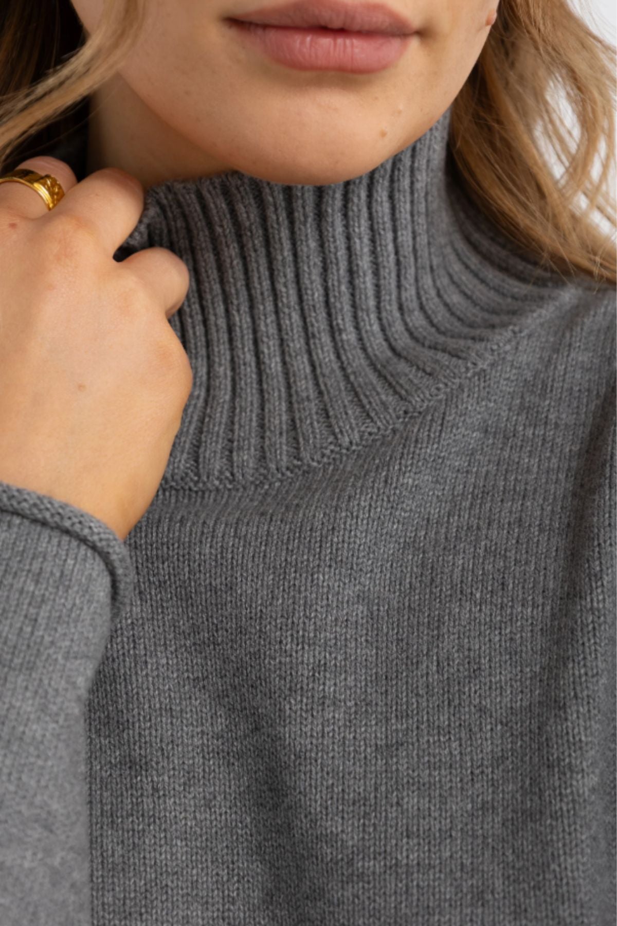 Close-up of a person wearing the ORTC Astrid Knit Sweater in grey, holding the collar. Wavy blonde hair and a gold ring enhance the elegant, cozy style of this cotton cashmere women's turtleneck.