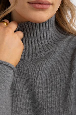 Close-up of a person wearing the ORTC Astrid Knit Sweater in grey, holding the collar. Wavy blonde hair and a gold ring enhance the elegant, cozy style of this cotton cashmere women's turtleneck.