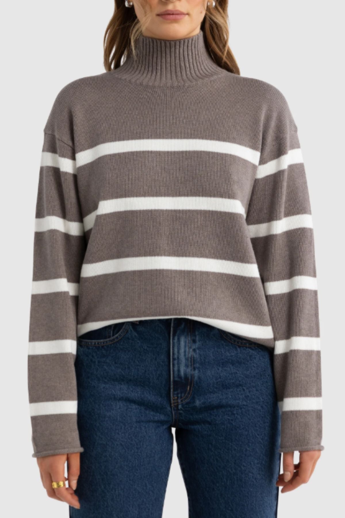 A woman wears the ORTC Astrid Knit Sweater, a taupe cotton-cashmere turtleneck with white stripes and long sleeves, styled with high-waisted blue jeans. The photo shows her from shoulders to hips against a plain background.