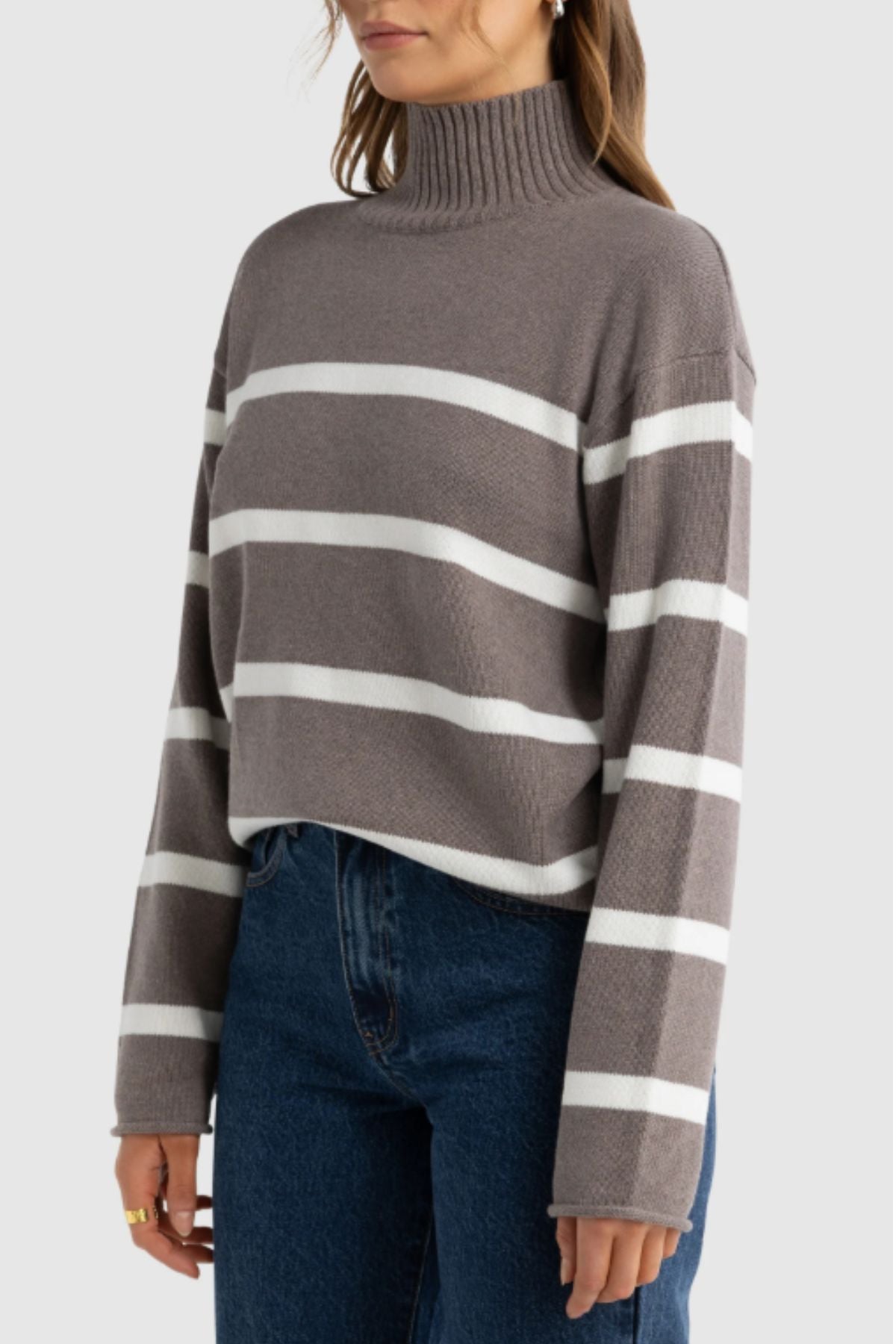 A woman wears the ORTC Astrid Knit Sweater—a gray turtleneck with white horizontal stripes in a soft cotton-cashmere blend—paired with blue jeans, standing against a plain light background.