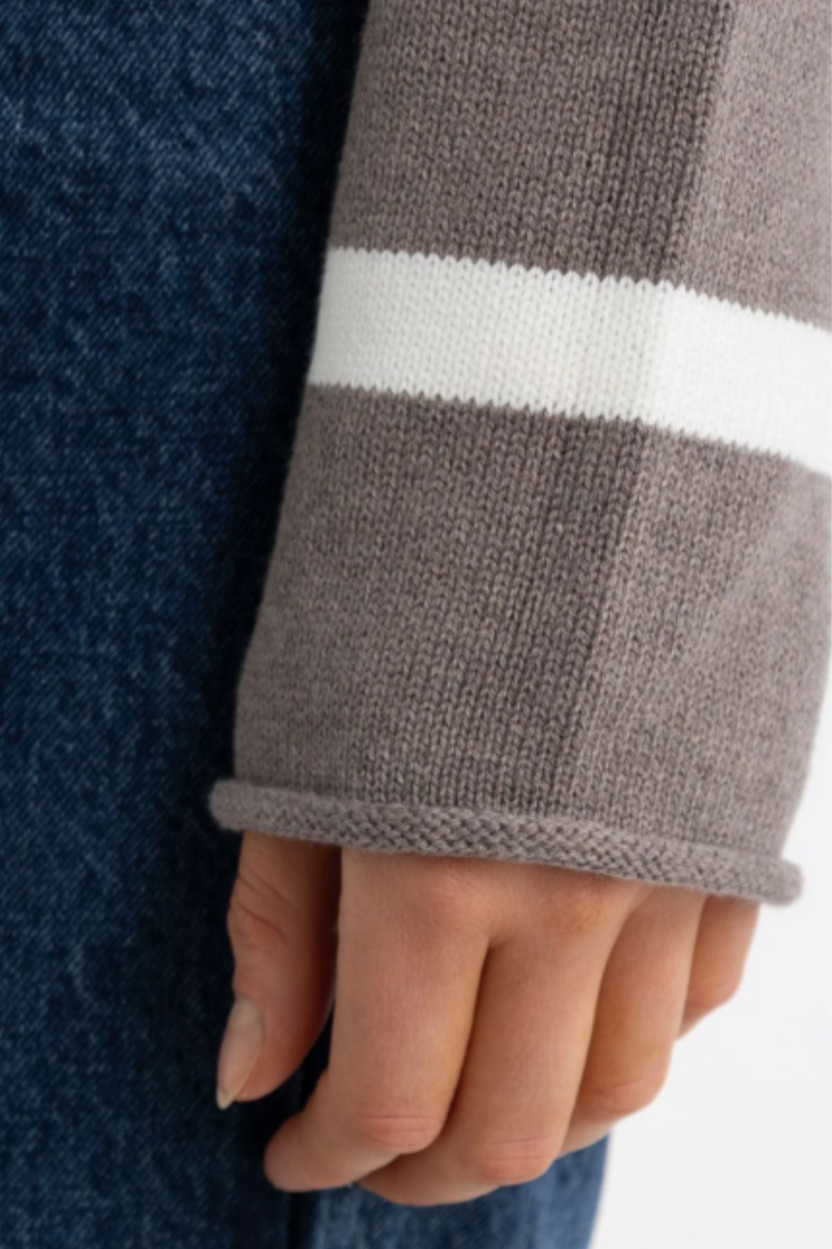 A close-up of a hand resting by their side, wearing the ORTC Astrid Knit Sweater in gray with a white sleeve stripe and blue denim jeans.
