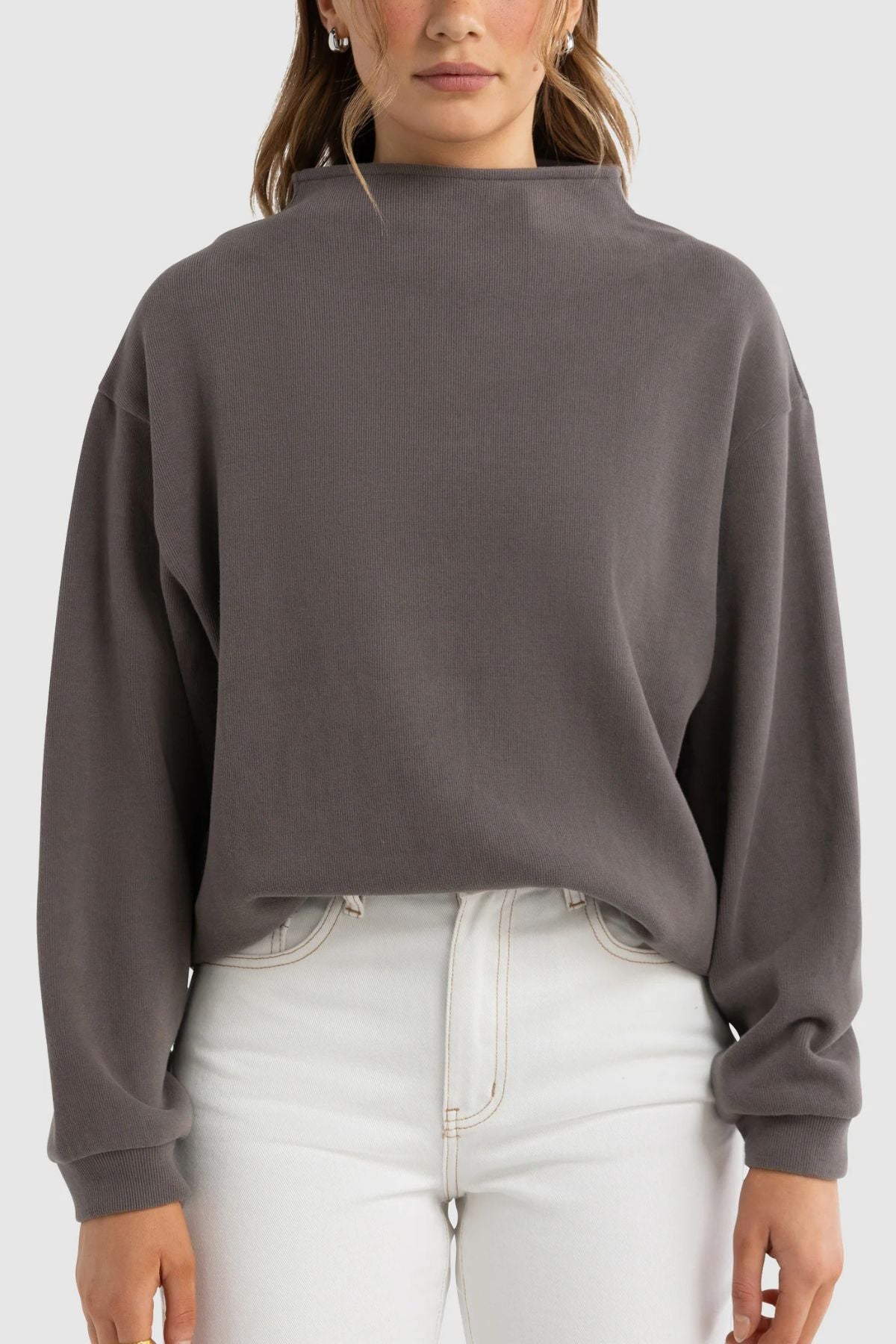 A woman stands facing forward against a plain background, wearing the ORTC Ava High Neck Jumper in dark gray with long sleeves and high-waisted white jeans.