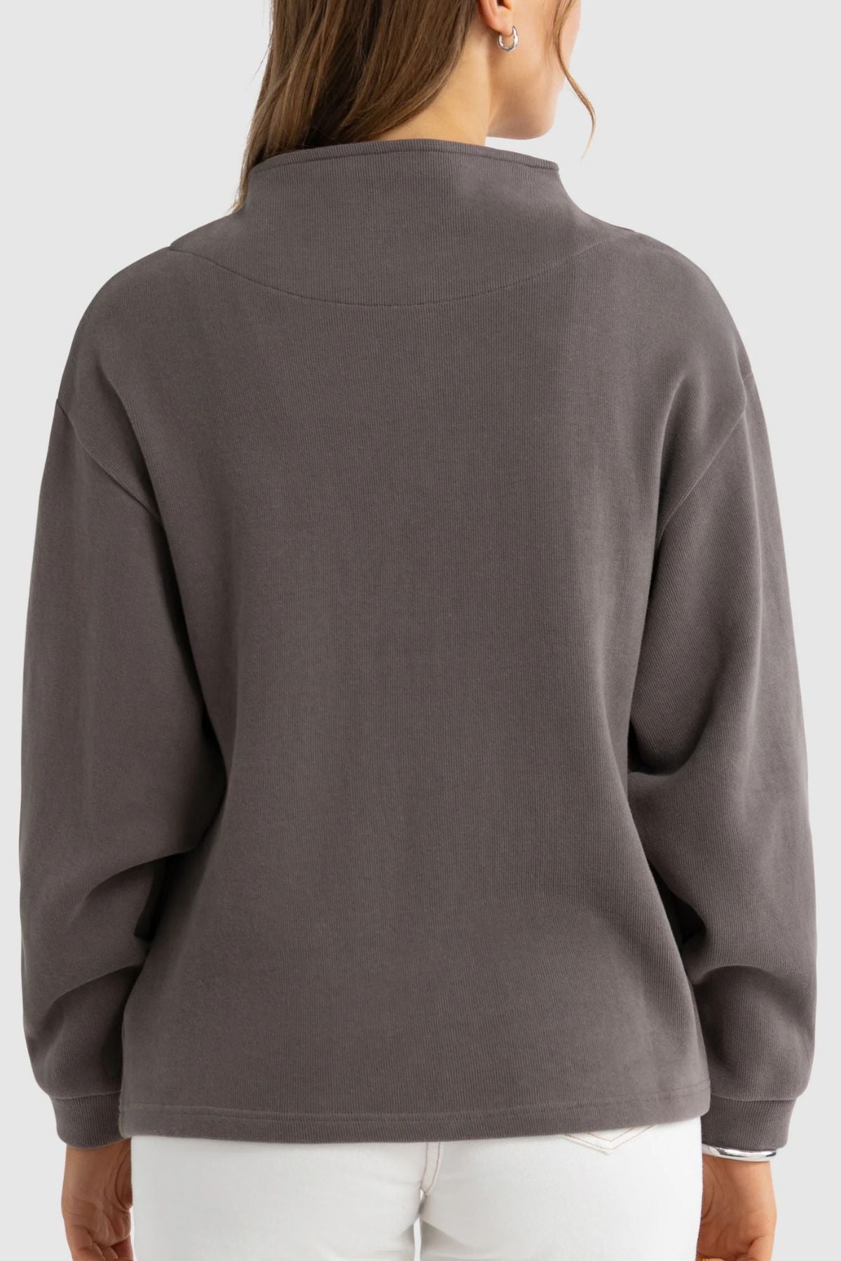 A woman with long brown hair is seen from the back, wearing the ORTC Ava High Neck Jumper in dark gray paired with white pants against a plain light background.
