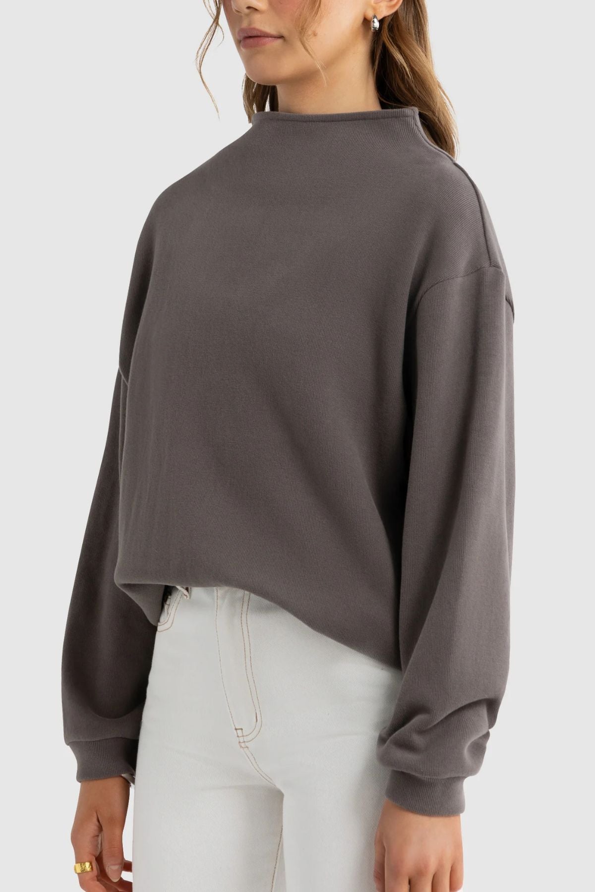 A woman wearing the ORTC Ava High Neck Jumper—a relaxed-fit, long-sleeved gray top—paired with high-waisted white jeans, standing against a plain light background.
