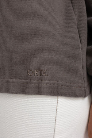 Close-up of the hem of the ORTC Ava High Neck Jumper in brown textured brushed cotton, featuring “ORTC” embroidered above light-colored pants.
