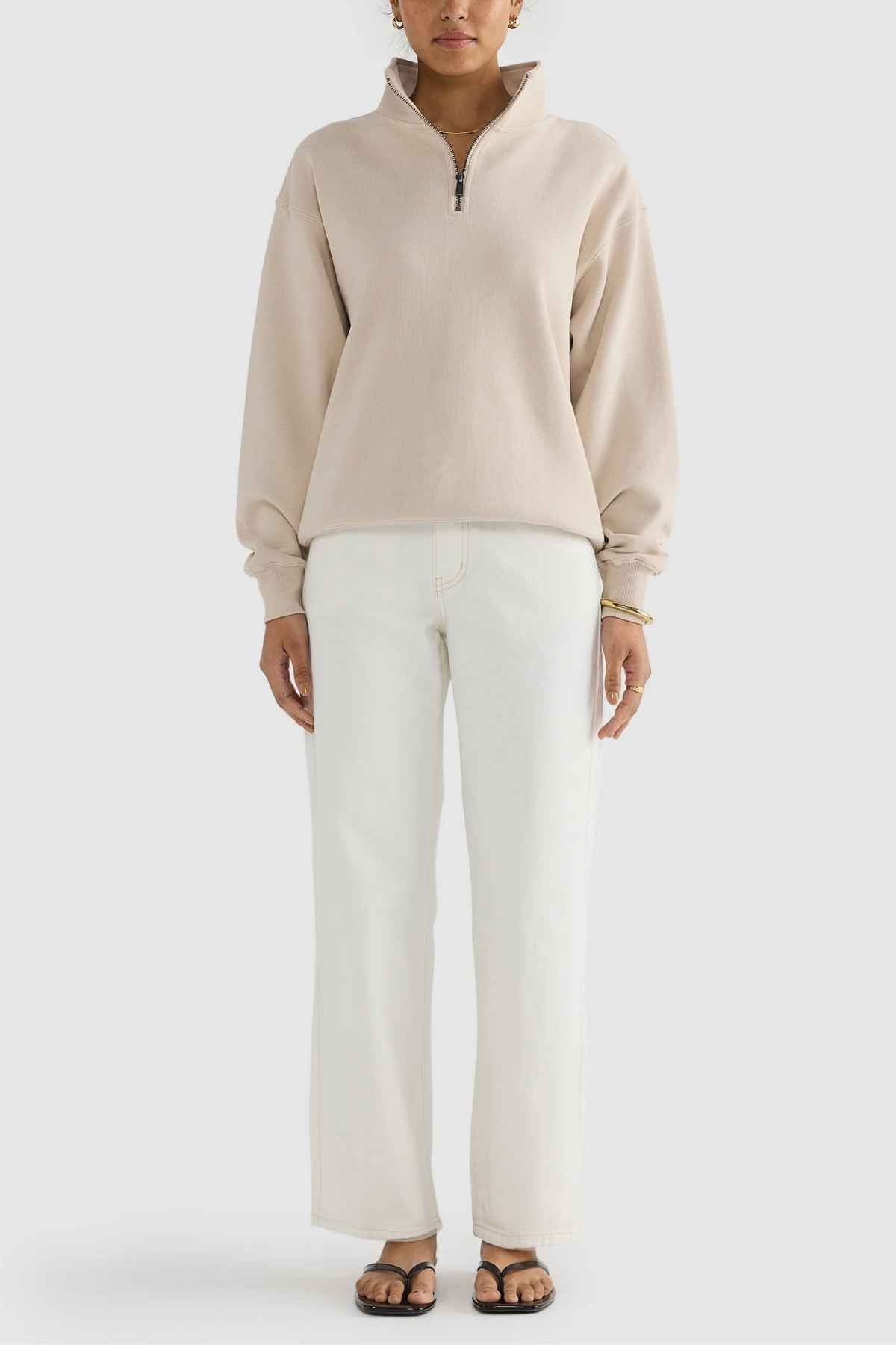 A person dressed in an ORTC Ava Ribbed Quarter Zip pullover in beige, paired with white wide-leg pants and black sandals, stands against a plain background with their hands by their sides. This chic ensemble is ideal for any capsule wardrobe.