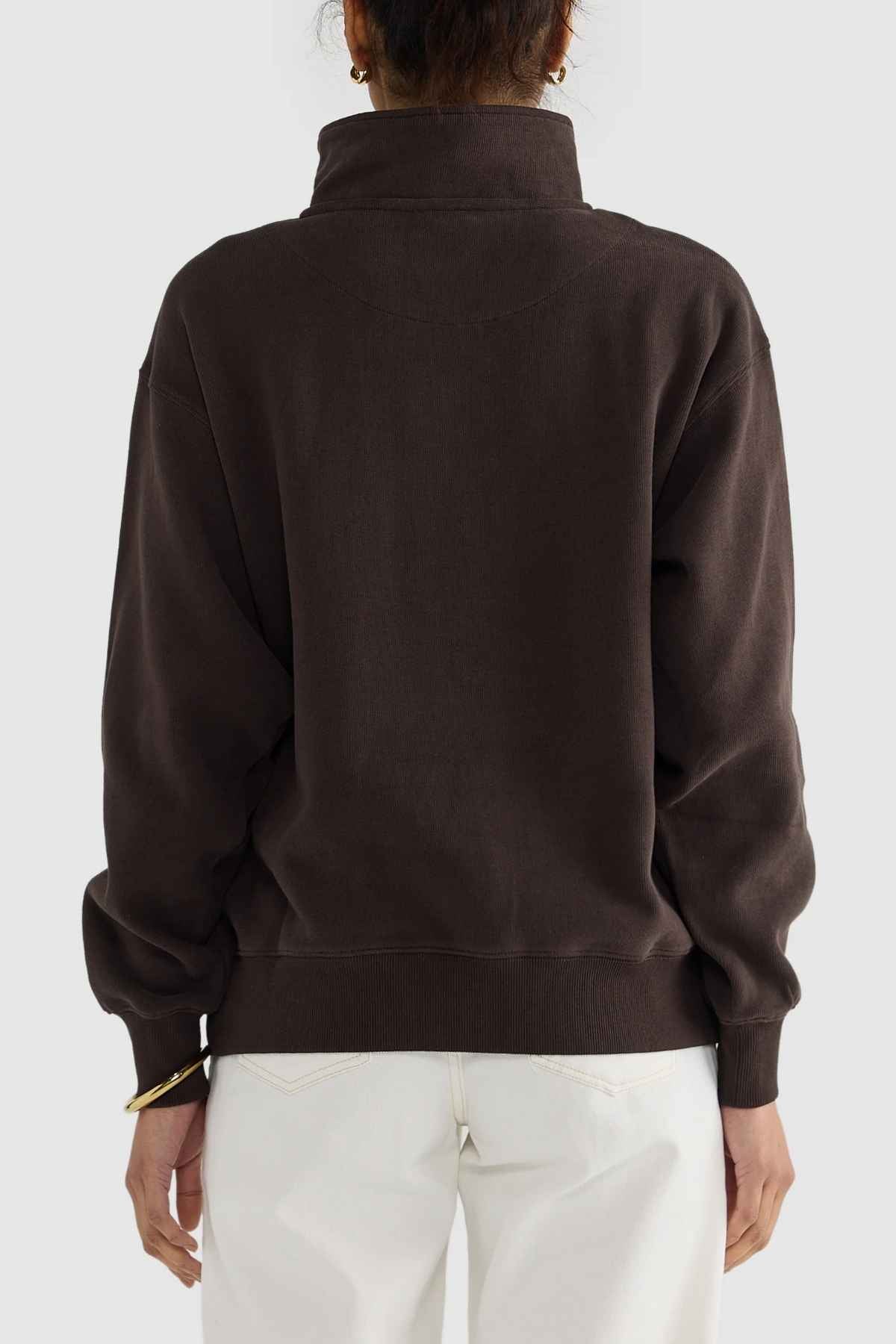 Rear view of a person wearing the ORTC Ava Ribbed Quarter Zip sweater in dark brown, paired with light-colored pants. The relaxed-fit sweater, crafted from brushed cotton, is complemented by the person's hair being tied up. The plain background accentuates the capsule wardrobe-inspired clothing.