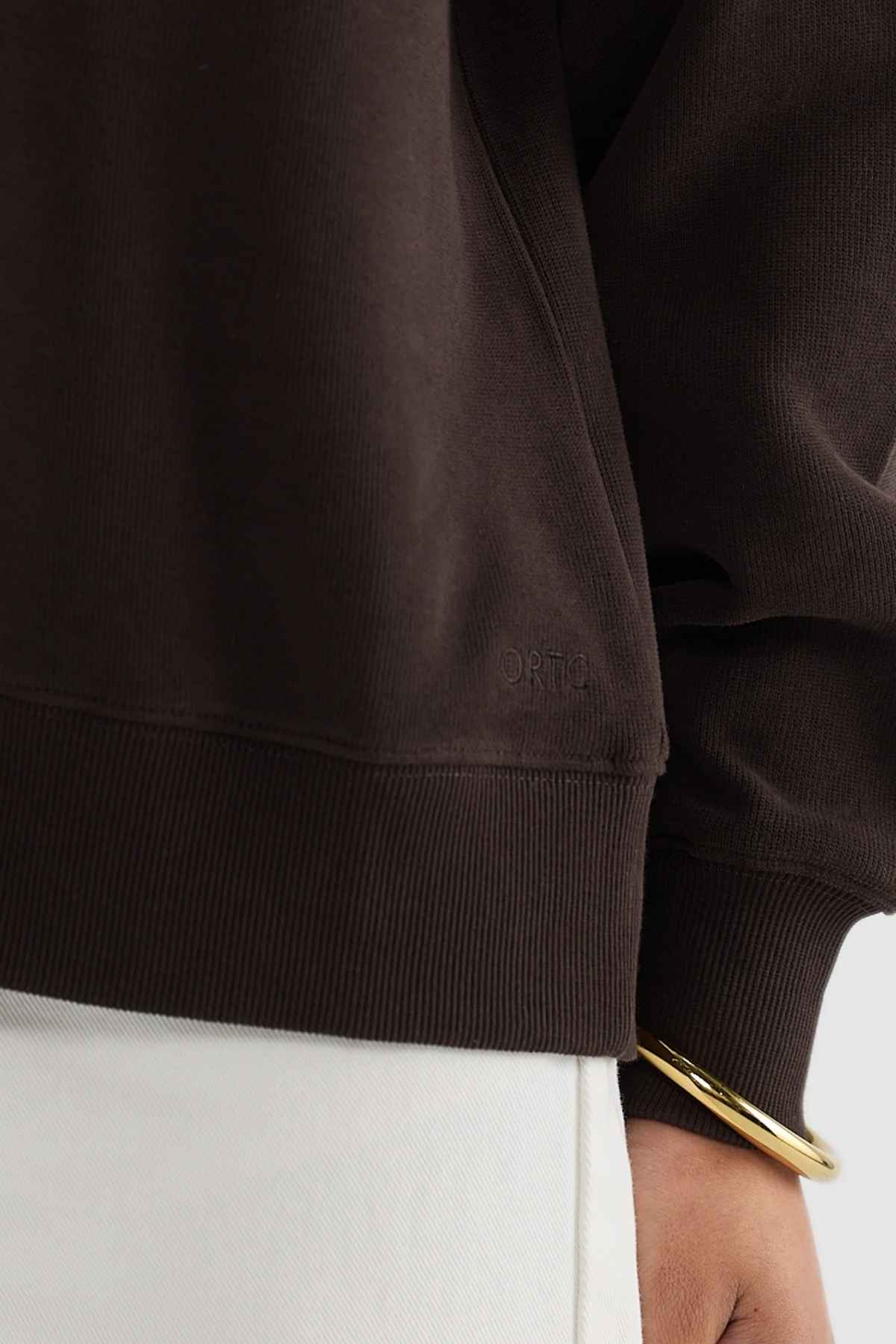 A close-up of a person's torso showcases them wearing the ORTC Ava Ribbed Quarter Zip, a dark brown brushed cotton sweatshirt with subtle text. The ensemble is completed with light-colored pants and a gold bangle on their wrist, highlighting the essence of a capsule wardrobe by emphasizing texture and color contrast.