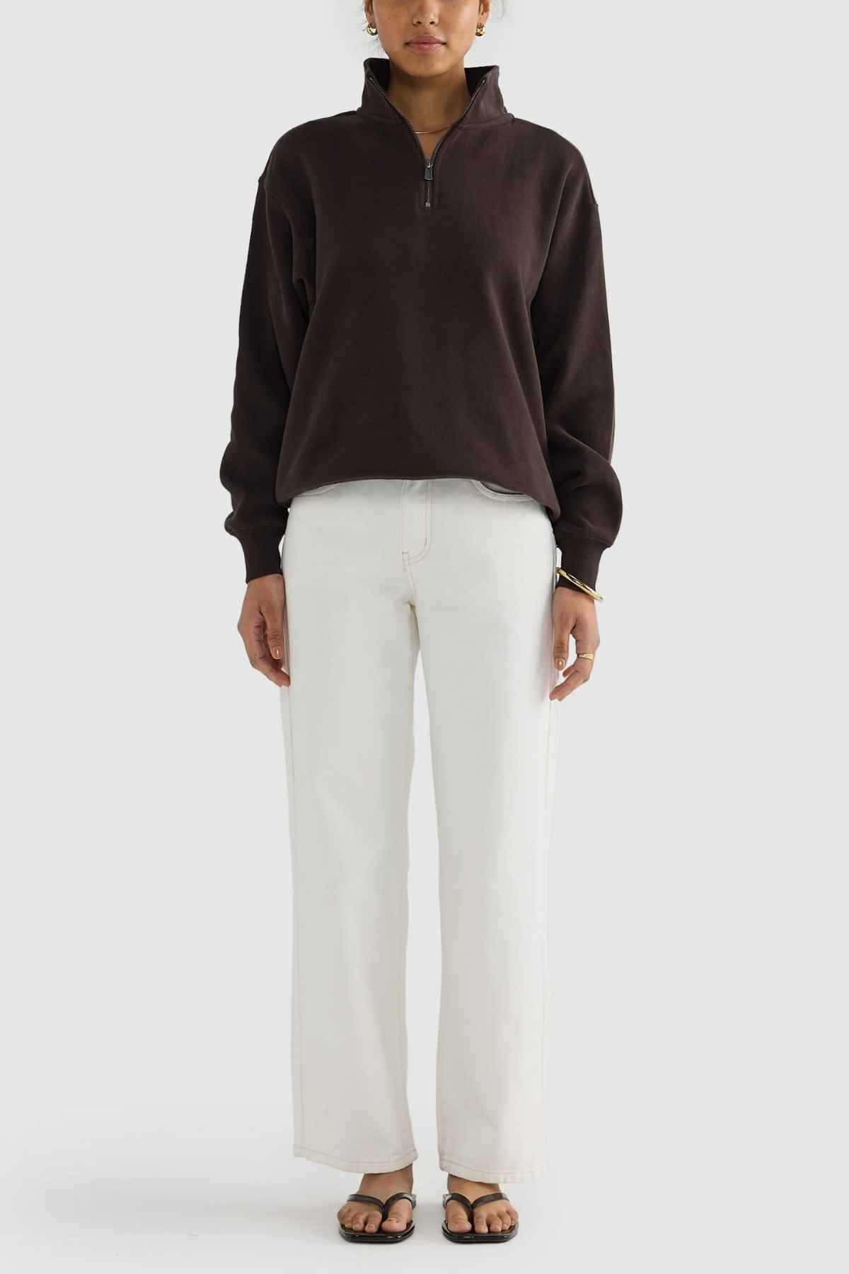 A person wearing a dark brown ORTC Ava Ribbed Quarter Zip sweater and white wide-leg pants, stands against a plain light background. Their capsule wardrobe look is completed with black sandals, and they have their hands at their sides.