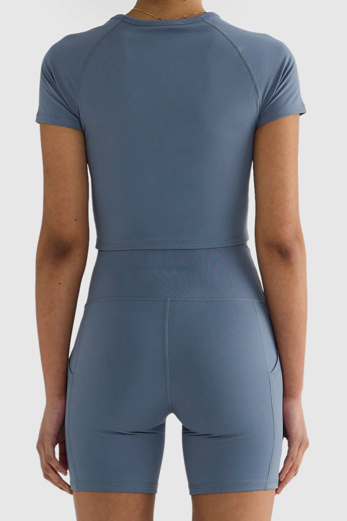 Rear view of a person wearing a matching blue athletic crop top and ORTC Eva 6 Inch Bike Shorts. The clothing is form-fitting and made from smooth, stretchy, recycled polyester fabric. The background is white.