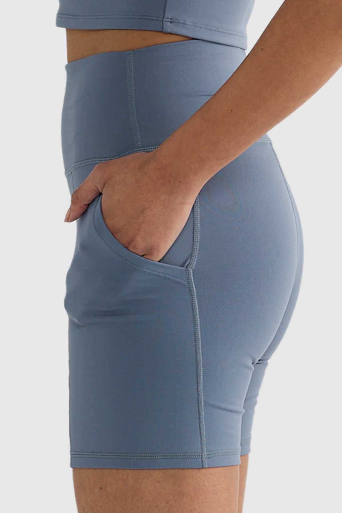 A person is sporting the ORTC Eva 6 Inch Bike Shorts in blue, with their hand tucked into the side pocket. The high-waisted shorts fit snugly and are crafted from recycled polyester, complemented by a matching top against a plain white background.