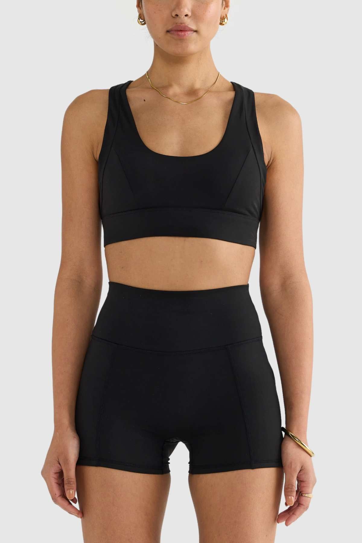 A person wearing a black ORTC Eva Crop Top, designed with brushed fabric, and high-waisted shorts is positioned against a simple light background. Their short hair complements the gold hoop earrings, necklace, and bracelet they are accessorized with, combining style and functionality effortlessly.
