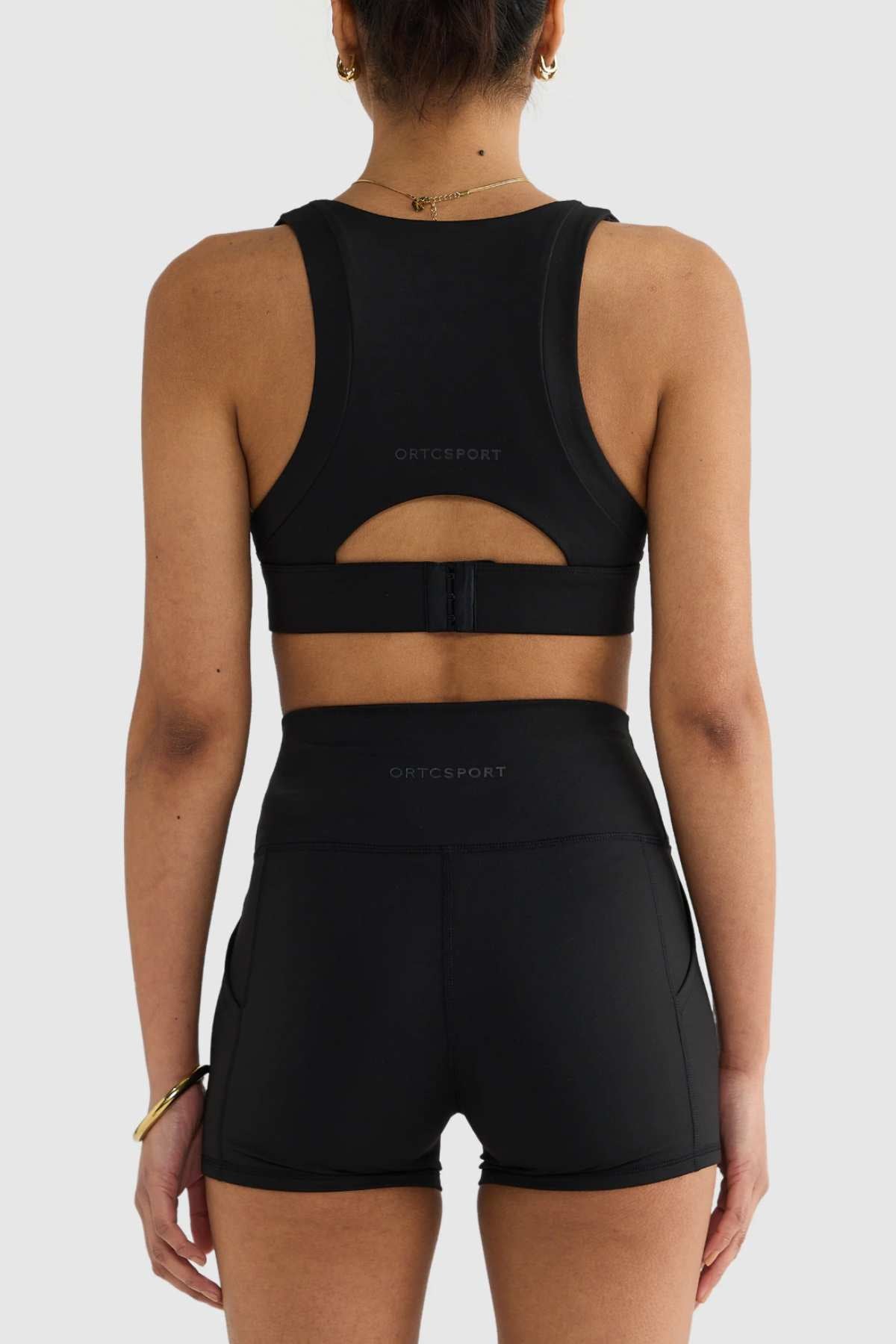 A person wearing an ORTC Eva Crop Top in black and high-waisted shorts is facing away from the camera. The outfit provides firm support and is complemented by bracelets, with their hair elegantly styled in a bun.